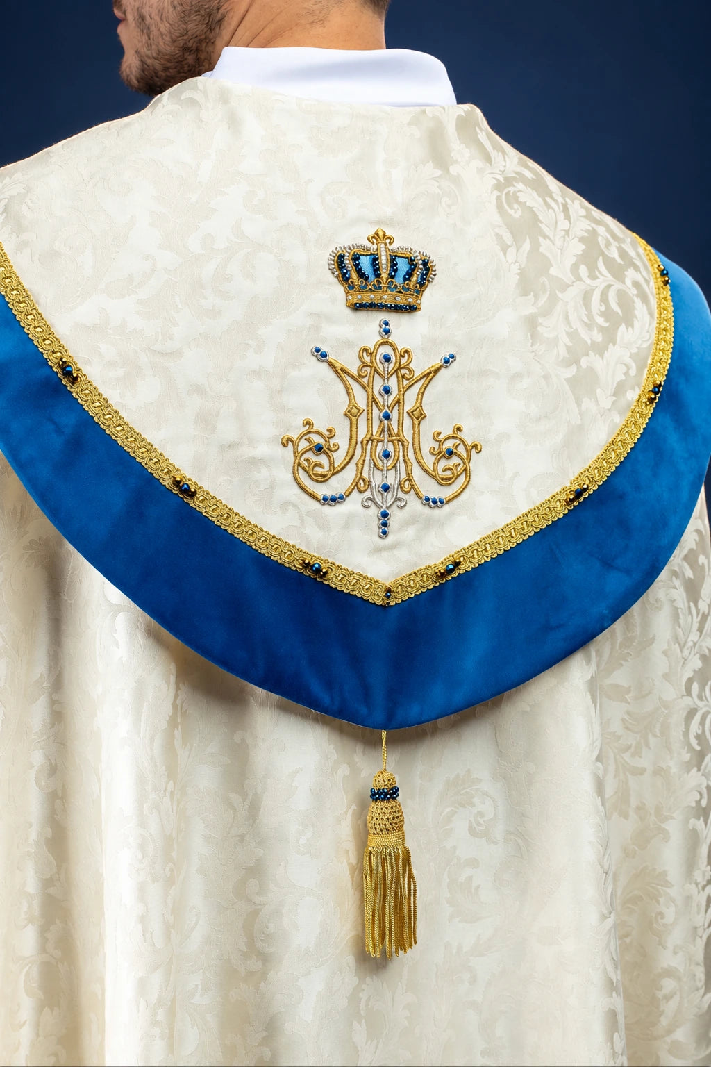 Back view of an ivory Marian cope with blue velvet orphrey, jeweled crown and monogram, gold tassel clasp