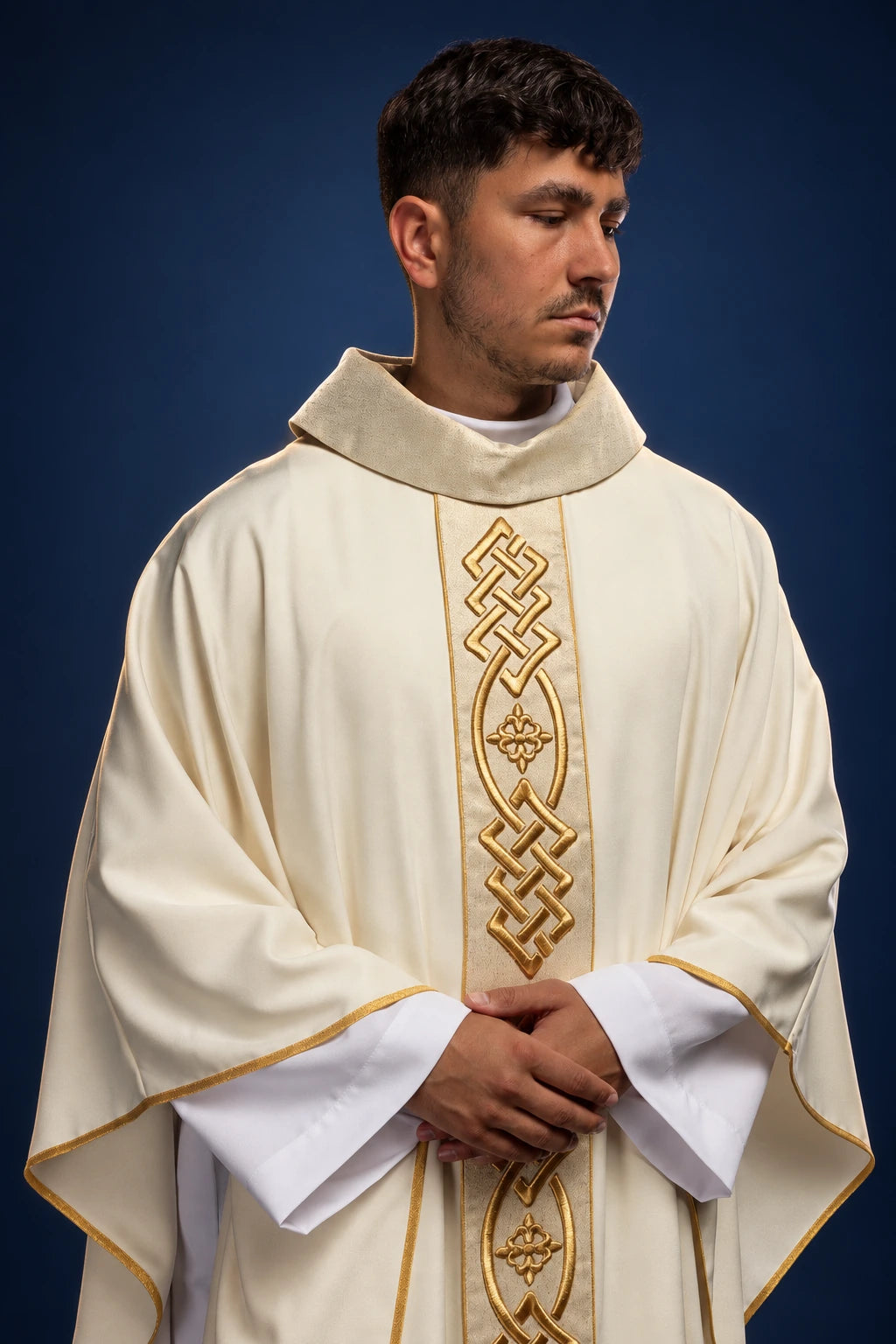 Front view of an ecru chasuble with gold embroidered cross and knot motif, worn over white sleeves