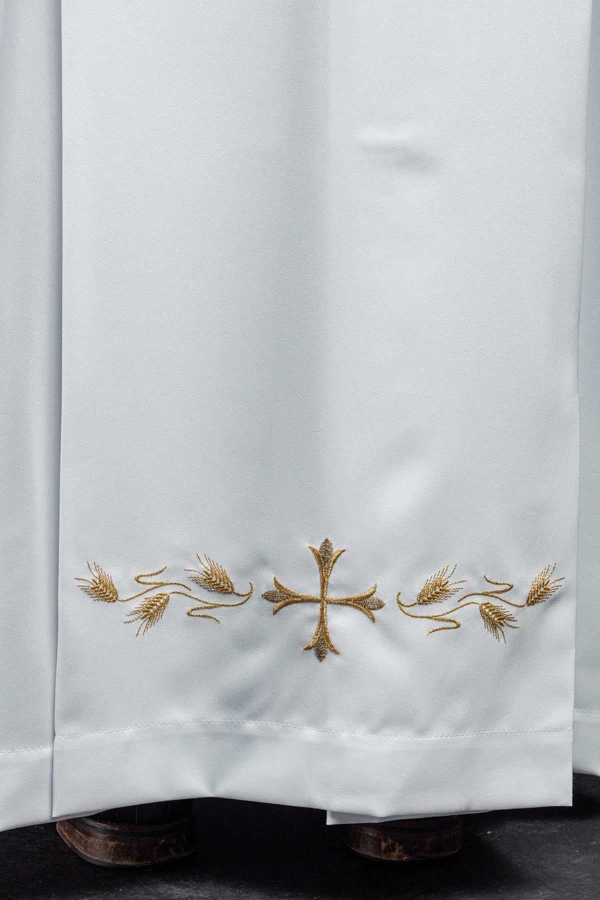 White priest alb close-up with gold cross and wheat embroidery at the hem