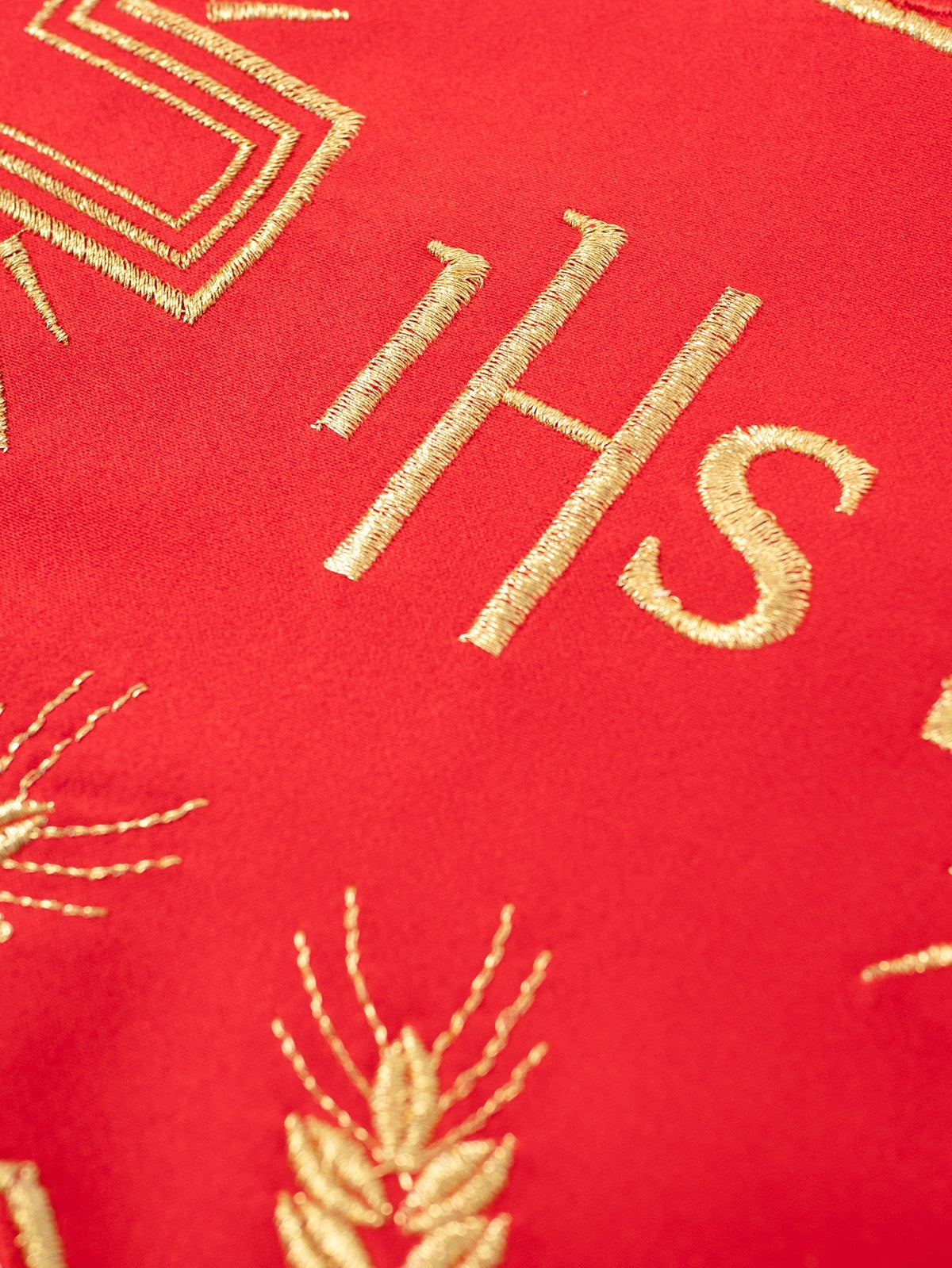 Red altar lace close-up showing embroidered gold IHS monogram, cross edge and wheat motif