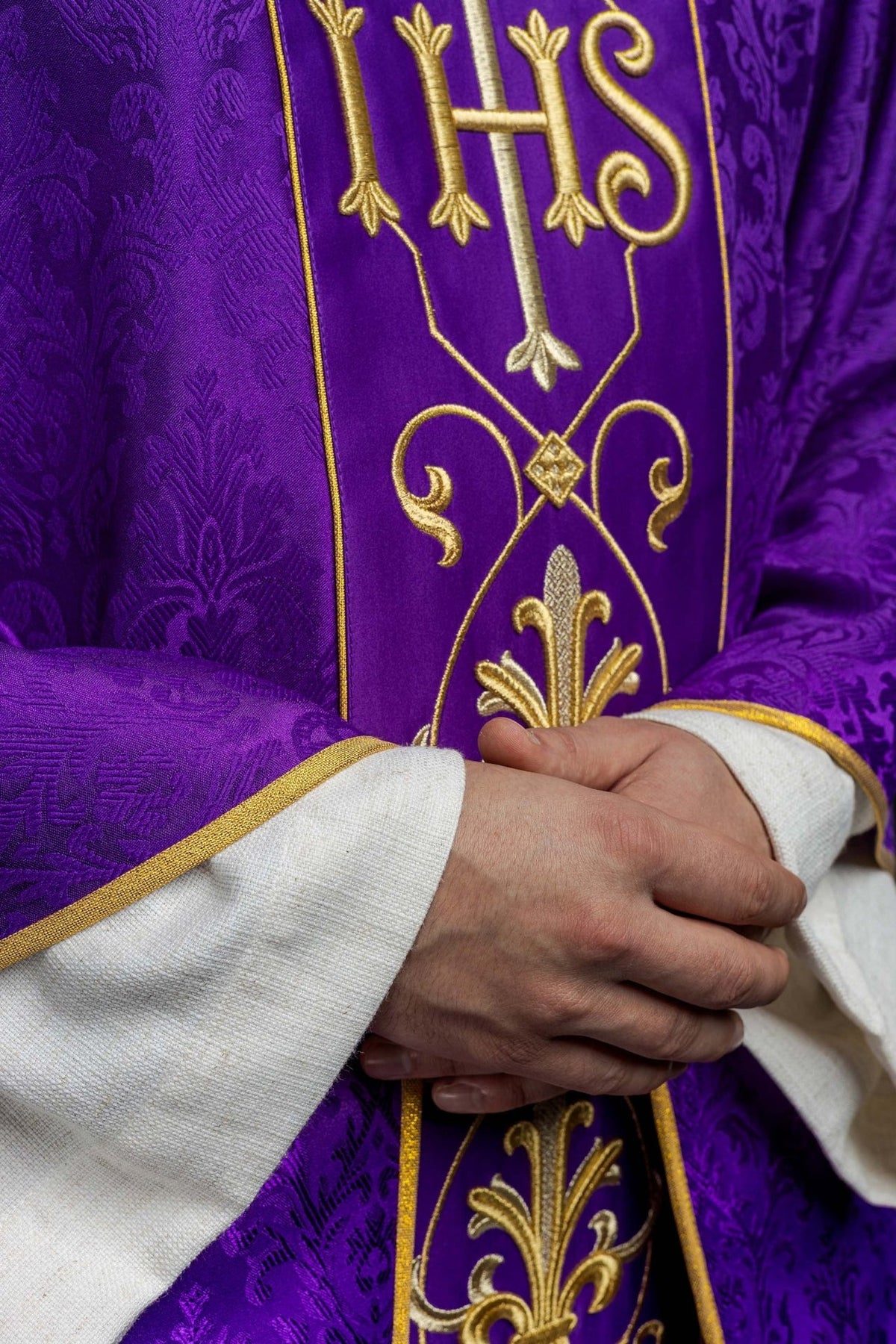 Purple chasuble close-up showing embroidered IHS panel and gold trim beside patterned fabric