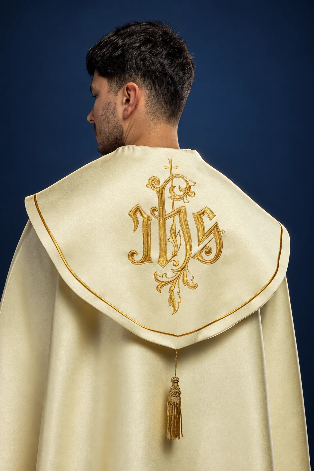 Back view of a cream cope with gold IHS monogram and ornate embroidered orphrey, tassel closure at the back neck