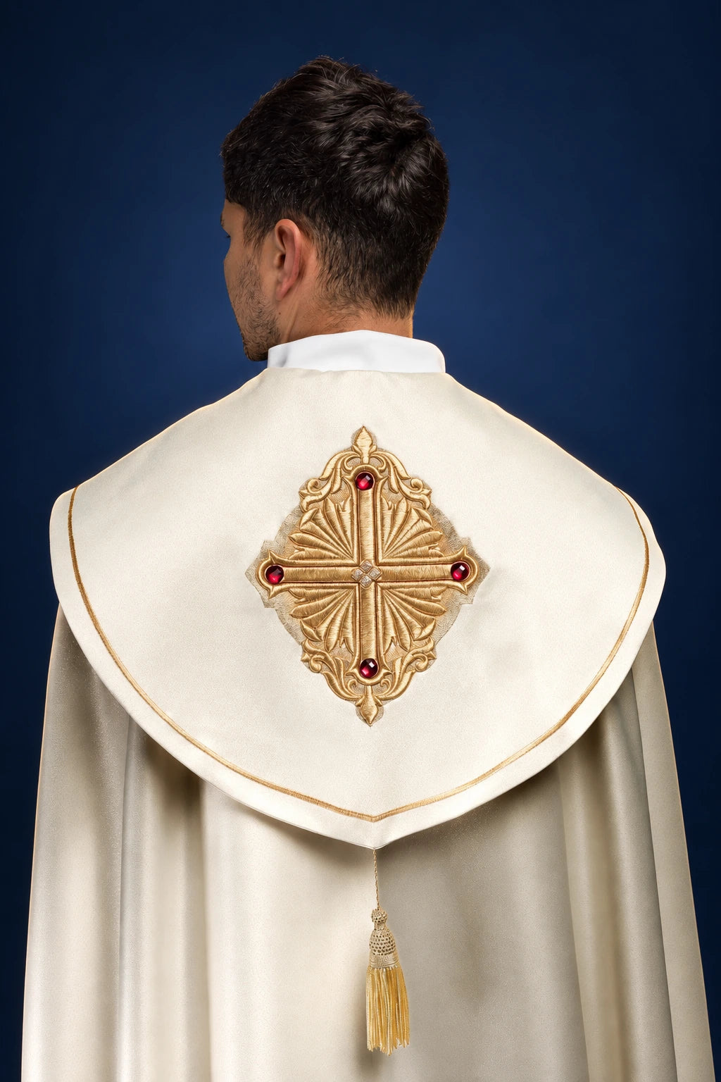 Back view of a cream cope with gold-trimmed hood and ornate cross orphrey with red jewel accents and tassel