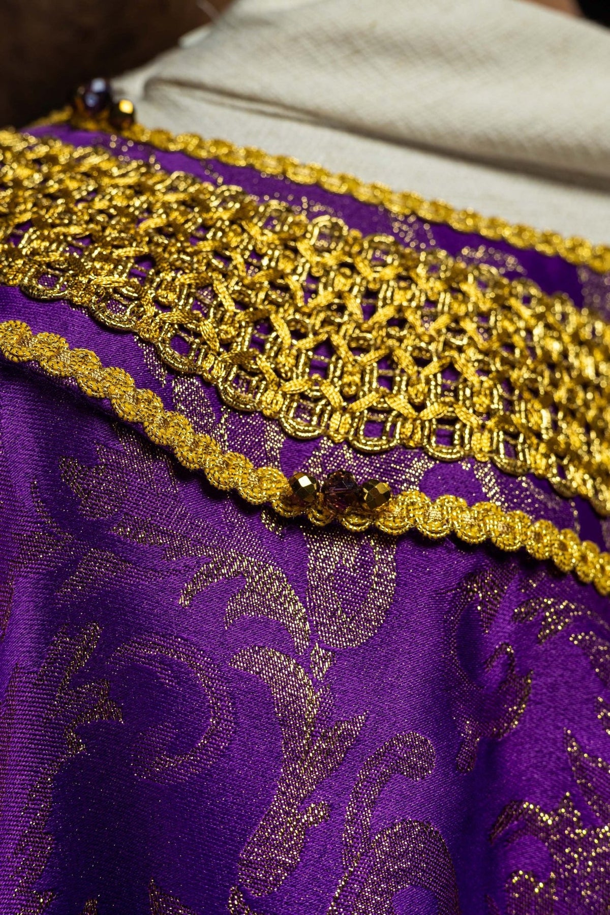 Purple chasuble close-up showing gold embroidery, trim, natural stones and jacquard fabric