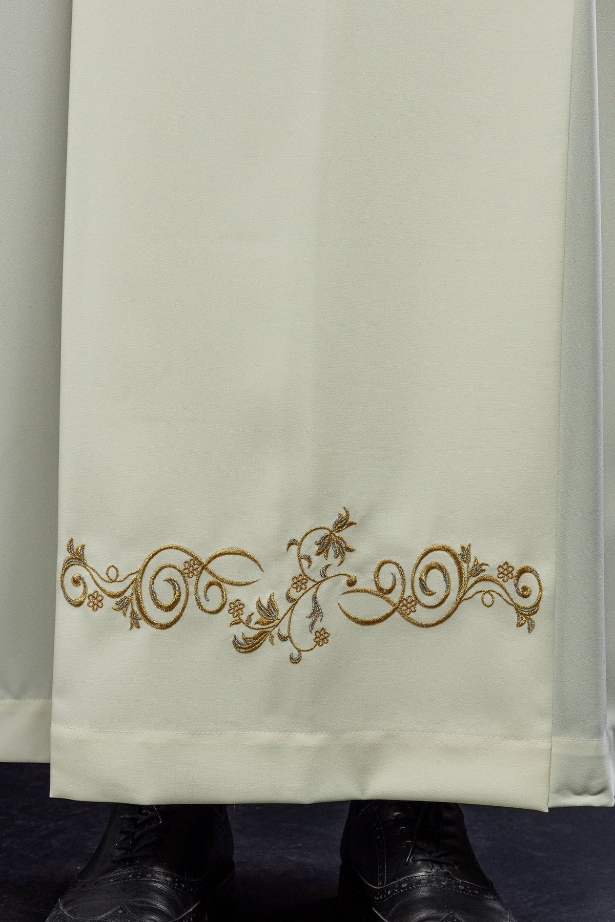 Ecru priest alb close-up showing gold decorative embroidery along the lower front hem
