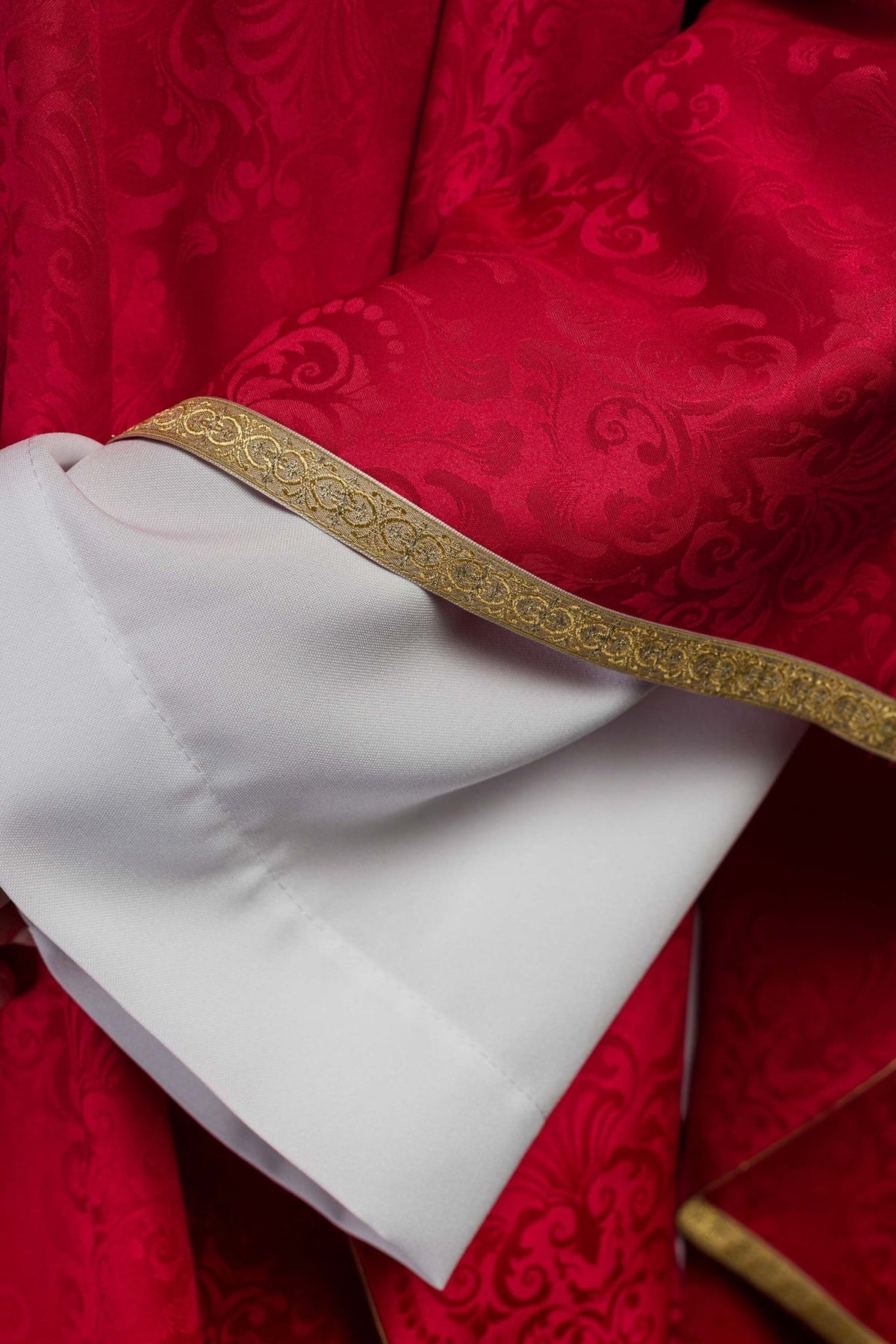 Red chasuble close-up showing jacquard fabric and gold trim over white sleeves