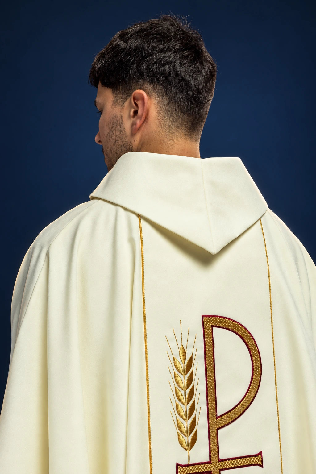 Rear view of an ecru chasuble with hooded back, gold piping, and embroidered wheat beside a Chi-Rho symbol