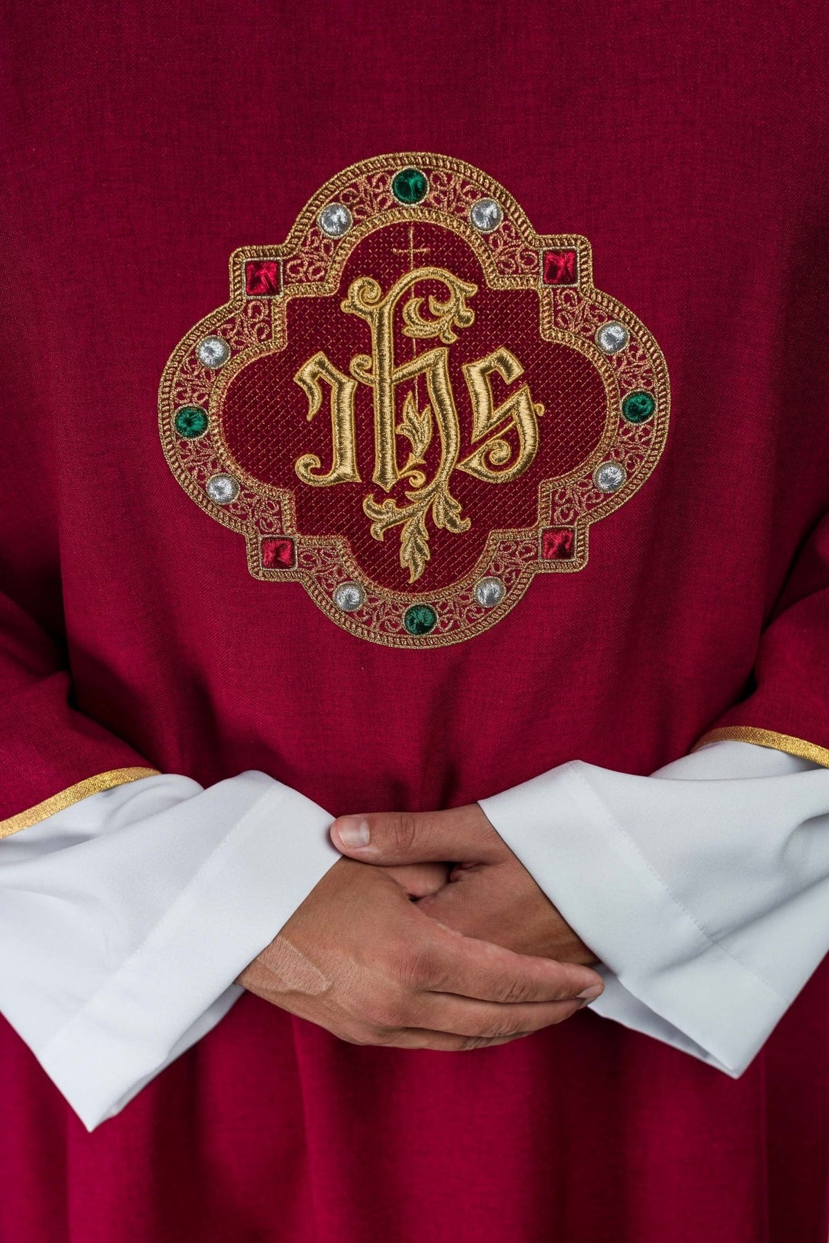 Red dalmatic close-up showing embroidered gold IHS symbol with green and silver details