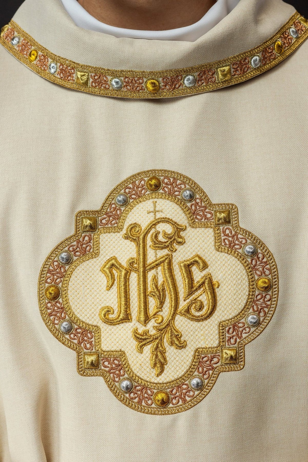 Ecru dalmatic close-up showing embroidered IHS symbol and ornate collar trim