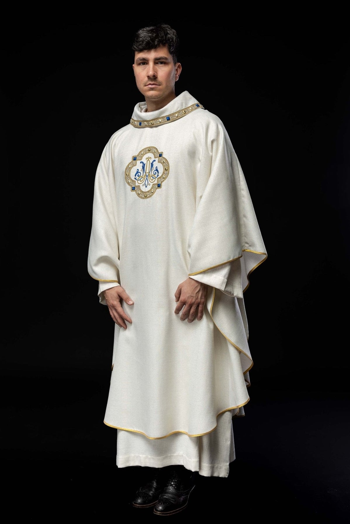 White liturgical chasuble worn, showing the embroidered Marian emblem and gold edging